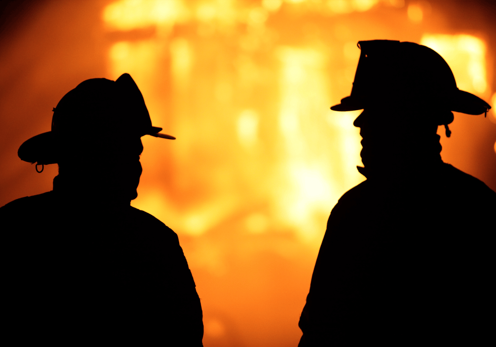 two firefighters stand silhouetted in front of a house fire