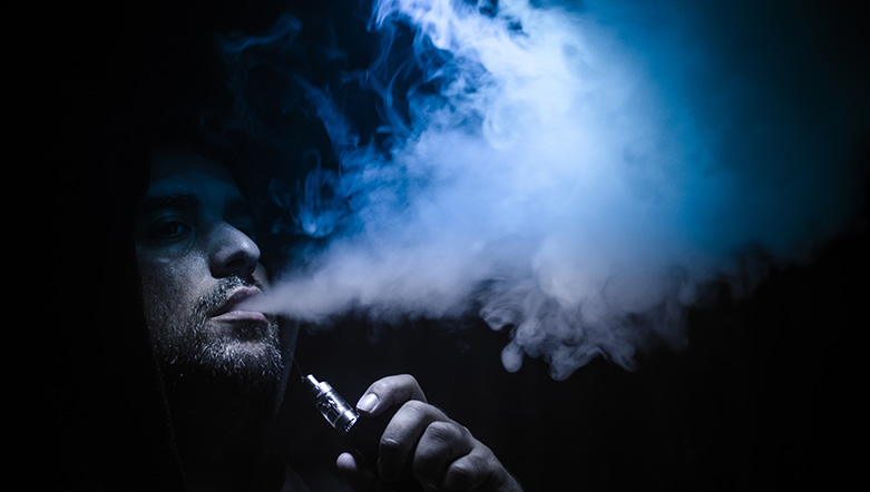 Man in dark hood blowing cloud of smoke from his vape